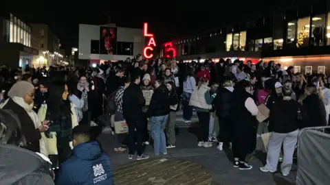 Hundreds of people dressed for the cold stand in a square on the university campus many holding banners 