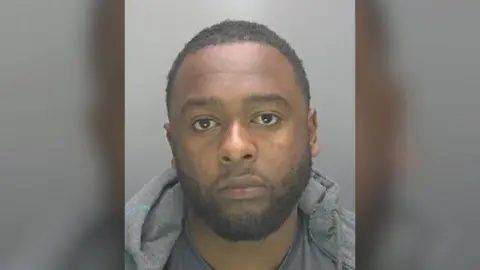 Cambridgeshire Police Custody photo of Nabil Aziz against a plain grey background. He has dark hair, brown eyes and a beard. He wears a grey hoodie, grey top and is looking directly at the camera. 