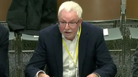 A man is sitting at a table and talking. He wears glasses and has a suit on with a white shirt and a yellow lanyard. A coat is sitting on an empty chair behind him.