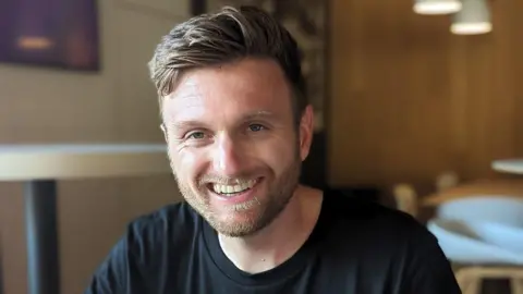 A head-and-shoulders image of Harry Oates. He is looking straight at the camera and smiling. He has light brown hair, longer at the top and shaved at the sides and a trimmed beard. He has blue eyes and is dressed in black t-shirt. The background is blurred.