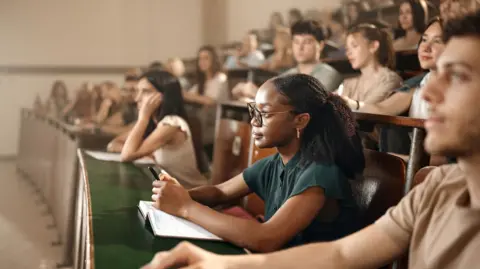 A young black woman wearing glasses is sitting at the front of a lecture hall, listening attentively - other students are visible around her