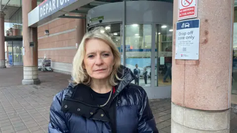 Elaine Miller - standing outside the departures area of Jersey Airport wearing a navy puffer coat.
