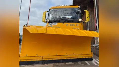 Suffolk Highways A close-up of the front of a bright yellow gritter lorry, which shows its cab and snow plough. 