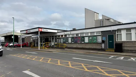 The exterior of Bletchley station which has a single-storey functional ticket office and waiting room, with a ramp and yellow handrail. There is a wooden portico with a station sign around the top. There are posters along the wall, and a single brown door to the right. There is doubled-door entrance under the portico. There is a strip of yellow "no waiting" paint on the roadway, which has white direction arrows on both sides.