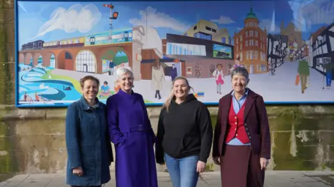 A group of women stand in front of a large rectangular mural. The mural features colourful depictions of local landmarks and buildings.