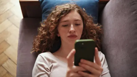 Getty  A teenager lies on a grey sofa looking at her phone, which has a green case. She has curly red hair and freckles, and wears a cream top.