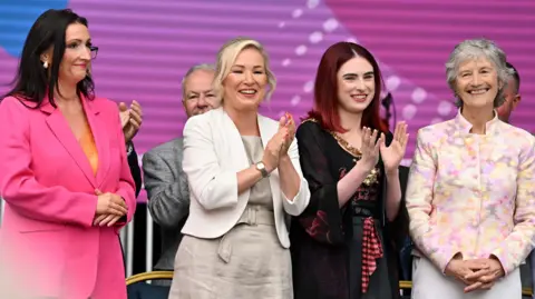PA Media Four women are standing together. The two on the outside have their hands clasped. The two in the middle are clapping. All are smiling. Other people and a large screen and chairs are in the background. 