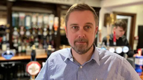 Andrew Sinclair/BBC Matthew Allum with a beard and open necked blue shirt stands at his bar. 