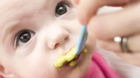 A stock picture of a baby's face with brown eyes looking up as they are spoon fed a liquid substance from a spoon.