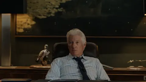 Paramount+ Richard Gere sits on an office chair wearing a tie and blue stripe shirt 