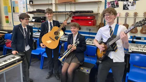 BBC Four schoolchildren, three boys and a girl, playing musical instruments in a music class. The boy on the far left has an electric piano, the boy on his right has an acoustic guitar, the girl in a middle has a trombone and the boy on the far right has an electric guitar