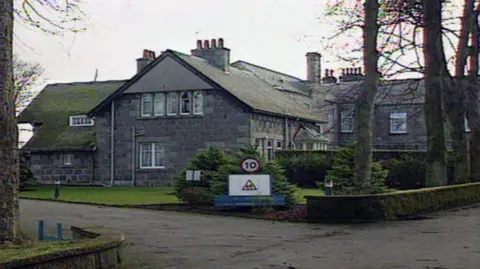 Entrance to Oakbank School with the main building behind a blue sign, grass, and trees.