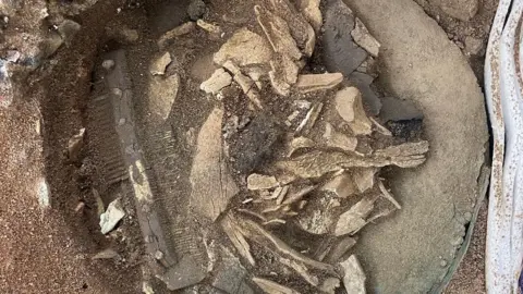 FAS Heritage/National Trust Human bone fragments as well as a comb are pictured in the earth, after being discovered. Soil and dirt surrounds the items. 