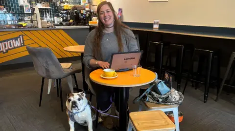 Vanessa Ward Photograph of Vanessa Ward, sat inside a coffee shop. She sits at a table with her 10-year-old bulldog, Bear. A coffee can be seen in front of Vanessa on a wooden table. She wears a grey polo shirt and smiles at the camera.