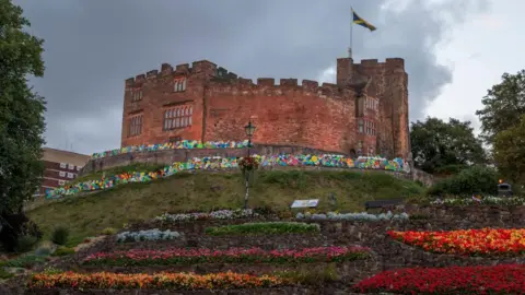 Getty Images A castle is on top of a hill surrounded by flowers and balloons. The sky is full of rain clouds.