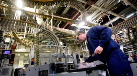 Getty Images Printer Gary Parslow repairs a machine at the Watford printing plant in 2010. Numerous conveyor tracks, containing newspapers, curve and loop through the space, creating a complex pattern across the ceiling.