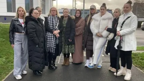 Ten women stand next to each other on a pavement outside a large building.