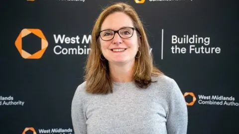Laura Shoaf stood against a WMCA backdrop, wearing a grey top and black glasses. She is smiling. 