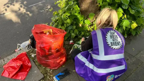 Brent Council A female council worker wearing a purple jacket, which says 'Don't mess with Brent' leans down to look through bags of rubbish
