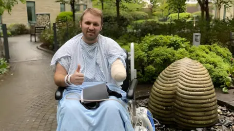 John Knight  John is pictured outside in a wheelchair, wearing a hospital gown with a blue blanket over his legs. He is smiling at the camera and giving a thumbs up. 