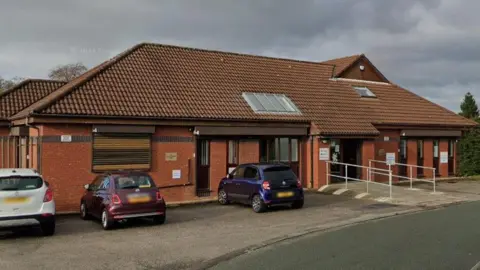 Google A single-storey brown brick building with three cars parked outside.