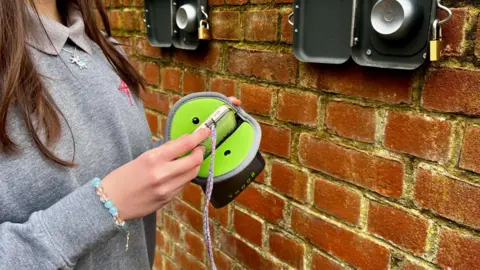 Sarah Smith/BBC A teenage girl puts her smartphone in a green protective bag to keep it magnetically locked for the day. There is a brick wall behind it. 