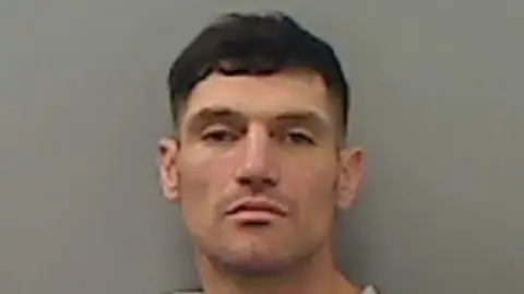 Mugshot of Aaron Thistlethwaite. He has a slight beard shadow and short dark hair. He may be attempting to look smouldering or aloof, but actually looks quite vacant.