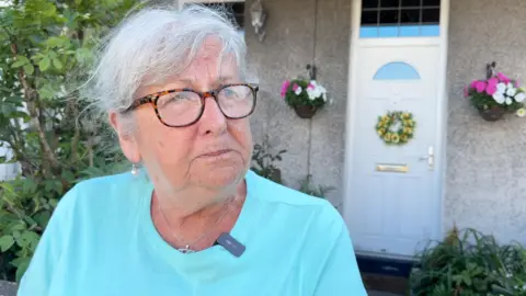 BBC Muriel Kelly has grey hair tied up and wears glasses. She has a light blue t-shirt on and is standing in her front garden. Behind her is a white front door with a flower wreath, and hanging baskets either side.