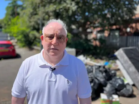A man with white hair and a white top is looking at the camera. Black bin bags are blurred on the photo on the pavement on the right.
