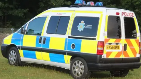 Cumbria Police Side view of a police van with blue and yellow markings, parked up in a grassy area. The rear side window has a Cumbria Constabulary logo.