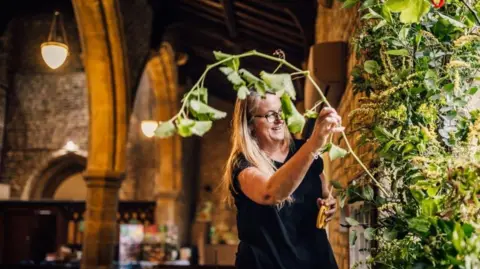 Pictured is Nicola Hill smiling with long blonde hair, wearing a black t-shirt and black round frame glasses. She is arranging flowers in a green plant flower display.