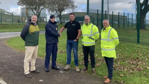 Royal Navy Commanding officer Capt Stuart Irwin wears a dark blue uniform and beret as he shakes the hand of Jason Thorpe who is wearing a black T-shirt and jeans. To the left of the Capt Irwin is Neil Howe, wearing an anorak and tan trousers, and to the right are Gareth Payne and Terry Grimes who are both wearing high-viz jackets and dark coloured trousers. They stand in front of a section of the perimeter fence and there are buildings in the background.