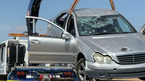 Cambs Police A large silver car is being winched on to a recovery truck. The car has a smashed front windscreen and both the driver and passenger doors are open