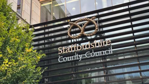 Staffordshire County Council's logo is on the side of a glass building. There is a tree in the foreground.