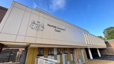 Martin Giles/BBC The outside of Norfolk Coroner's Court. The building has a sign in black writing which says Norfolk Coroner's Court. There is also trees and blue sky in the picture.