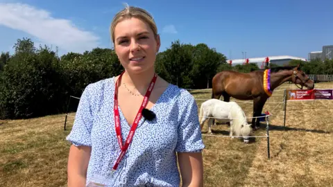 Lilly Pinchin, wearing a blue and white top, in a field with two horses behind her.