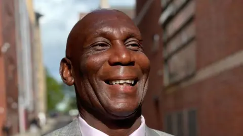 Uriah Rennie, a man wearing a grey jacket, lilac shirt and purple tie. He is smiling and appears to be walking down a narrow street between two large redbrick buildings.