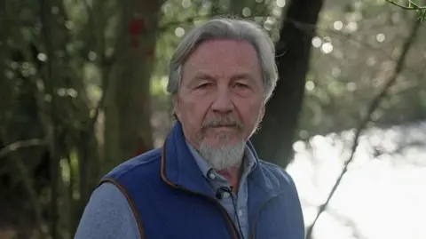 Steve Ormerod wears a blue gilet and a blue shirt. He has grey hair and a grey beard