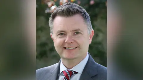 Royal School Armagh Graham Montgomery. He has shirt dark hair with grey patches, wearing a grey blazer, white shirt, and red striped tie.