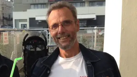 Peter Roscoe A man with brown hair, a beard and glasses wearing a white tshirt and black jacket standing on a balcony with a metal fence behind him