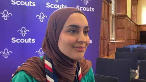 Ayesha Karim smiling at the camera. She's wearing a brown headscarf and a stripy scouts neckerchief. There's a purple scouts banner behind her