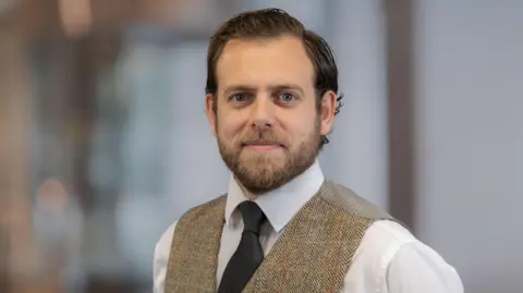 Savills Guernsey Stuart Leslie, a man with brown hair and a brown beard. He is wearing a white shirt, dark tie and a beige waistcoat. He is looking at the camera. The background is blurry.