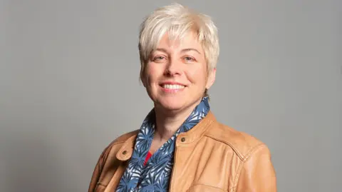Cámara de los Comunes Una mujer con cabello rubio corto, una chaqueta marrón claro y una bufanda azul y blanca, se para frente a un fondo gris y sonríe.