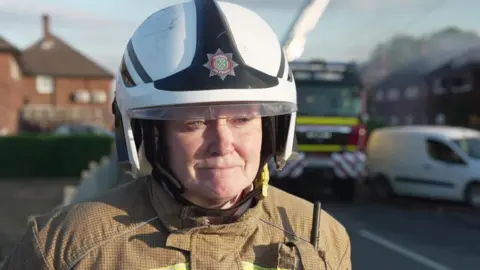 JOE WEIR / BBC A woman with a white fireservice helmet and fire uniform is standing in the street looking into a camera. Behind her is a fire engine, a row of cars and the remains of a burnt out house.