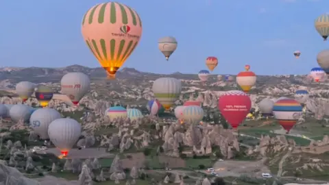Josh Kohler/Instagram A rocky landscape with unusual stacks of large pointy rocks over a large area. About 30 colourful hot air balloons rise from the scene, against a blue sky 