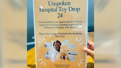 Atiq Rehman A hand holding up a poster advertising the drop off point, which reads "Unspoken Hospital Toy Drop 24", with a photo of a child holding a teddy bear