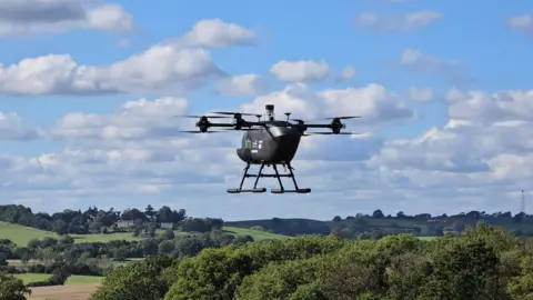 A prototype vertical take-off and landing aircraft with four separate rotors flying over fields and trees on a clear day. The aircraft looks like a helicopter but with four separate rotors instead of one big one.