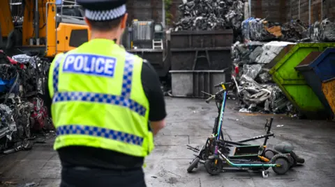 A police officer looks over a pile of confiscated electric scooters in a scrap yard with piles of crushed metal in skips in the background