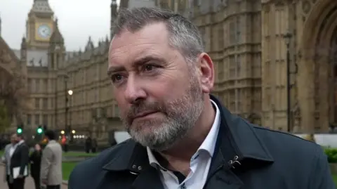 A still of Des Conroy outside Westminster. He is being interviewed. He is a middle aged man who has short, greying hair and a beard. He has a neutral expression on his face and is wearing a black coat with a white buttoned shirt.