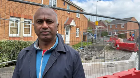 Sri Madadi, a man wearing a navy blue coat with a light blue fleece underneath, is standing by some metal fencing which surrounds the front of a house, with a pile of gravel and chunks of broken pavement in front of it. The houses are brick-built, with white window frames and door.
